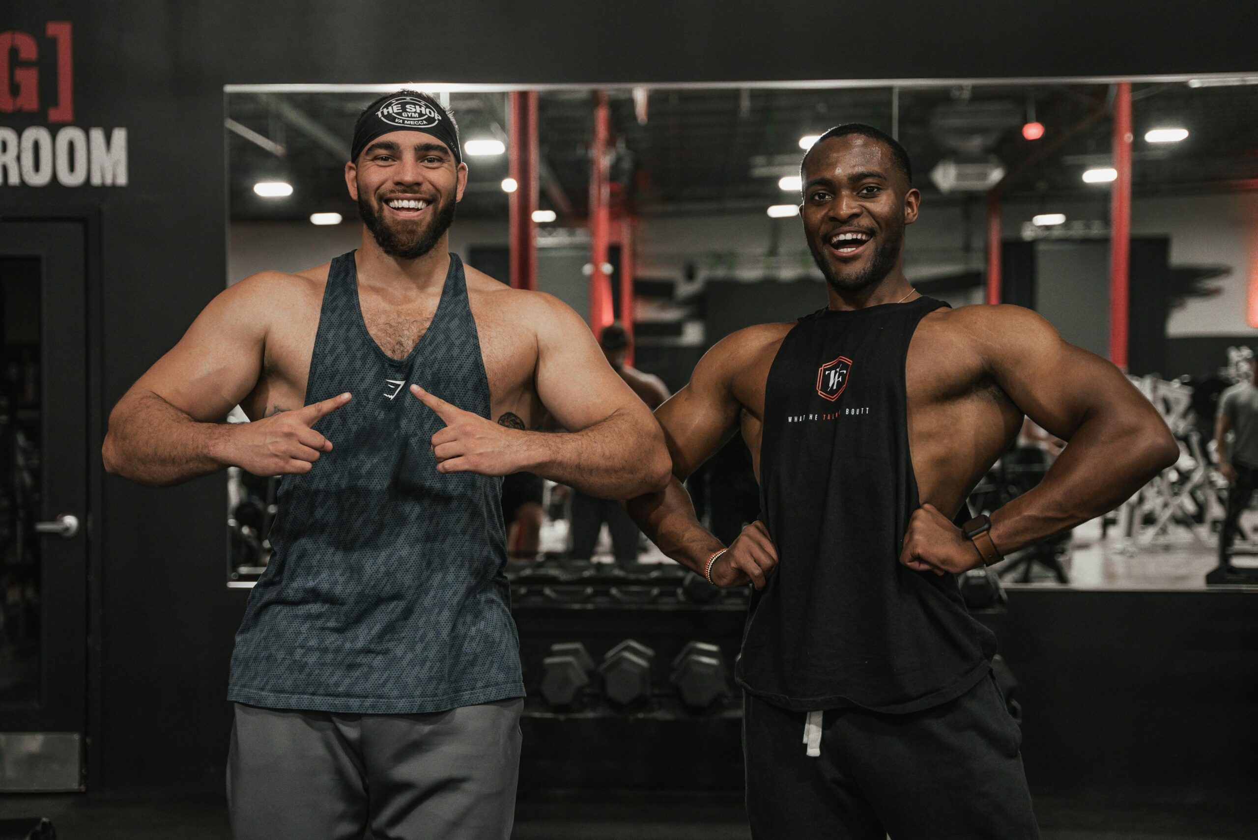 Two muscular men smiling while posing confidently in a gym environment.