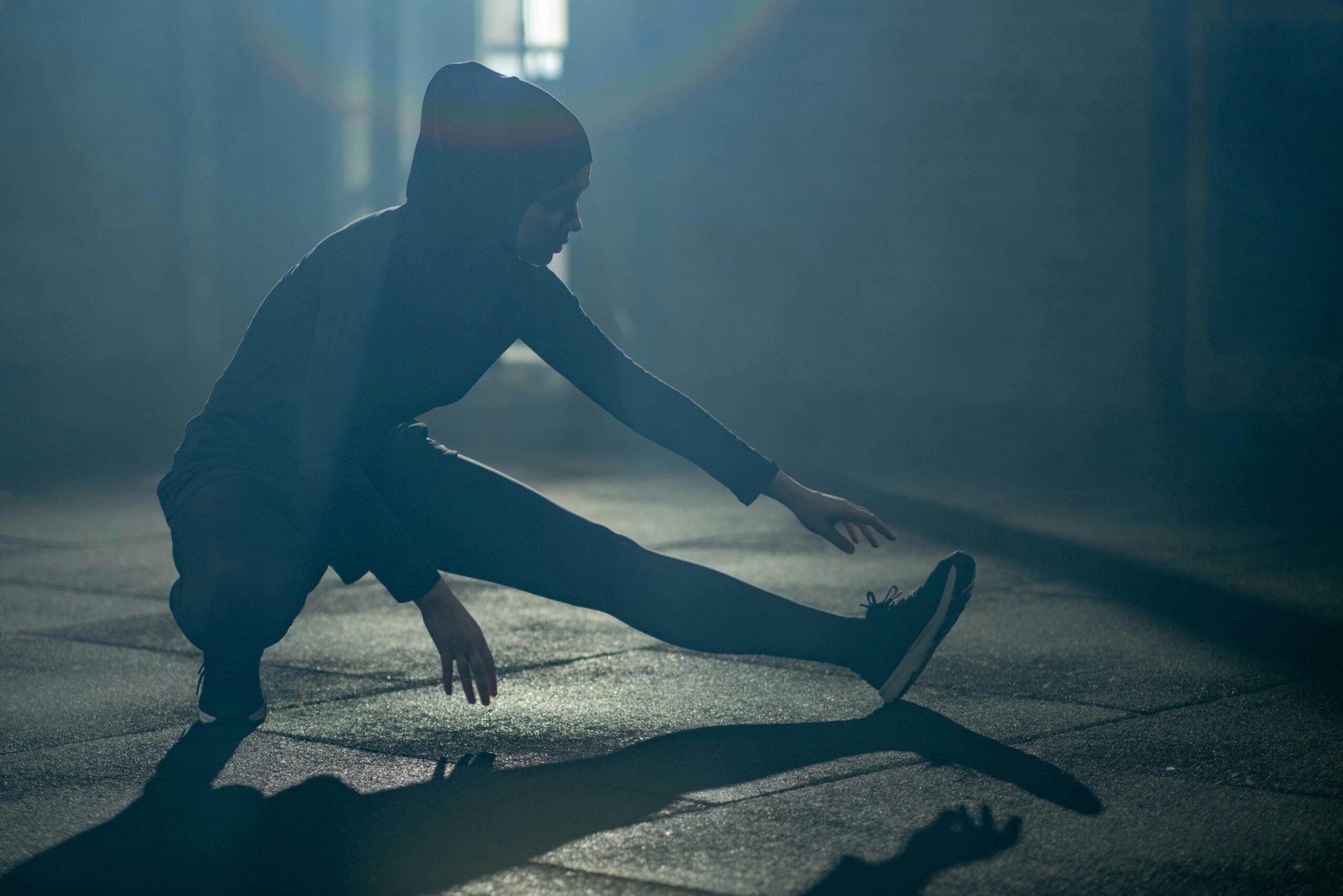A Muslim woman in activewear stretches indoors, bathed in dramatic light. Fitness and focus.