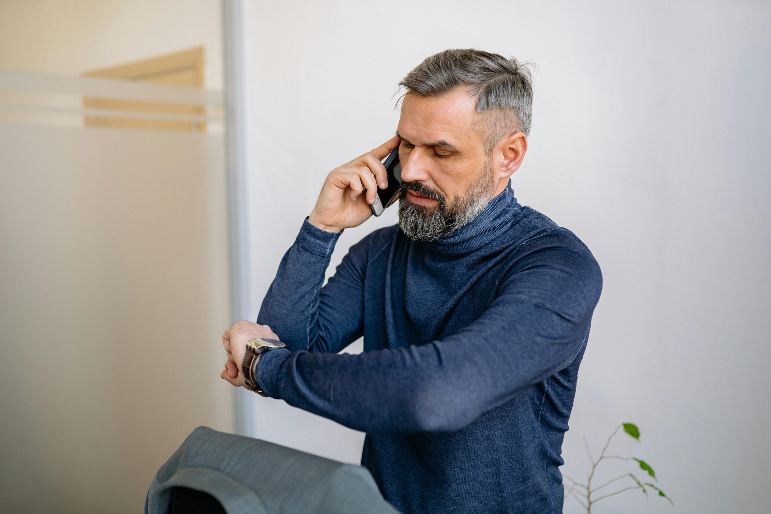 Man with beard speaking on phone while checking the time indoors.